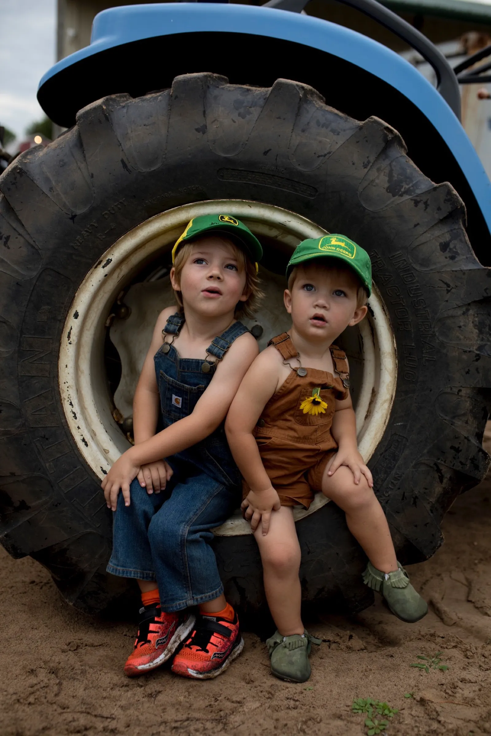 Two of Roger Marshall's grandsons in John Deere caps sitting by a tractor tire.